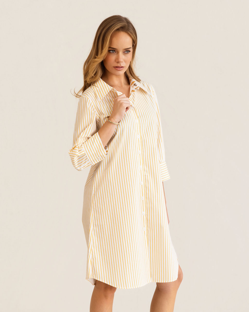 A woman modeling a yellow and white Kaleido cotton vertically striped button-down shirt dress with three-quarter sleeves, standing against a plain off-white studio background.
