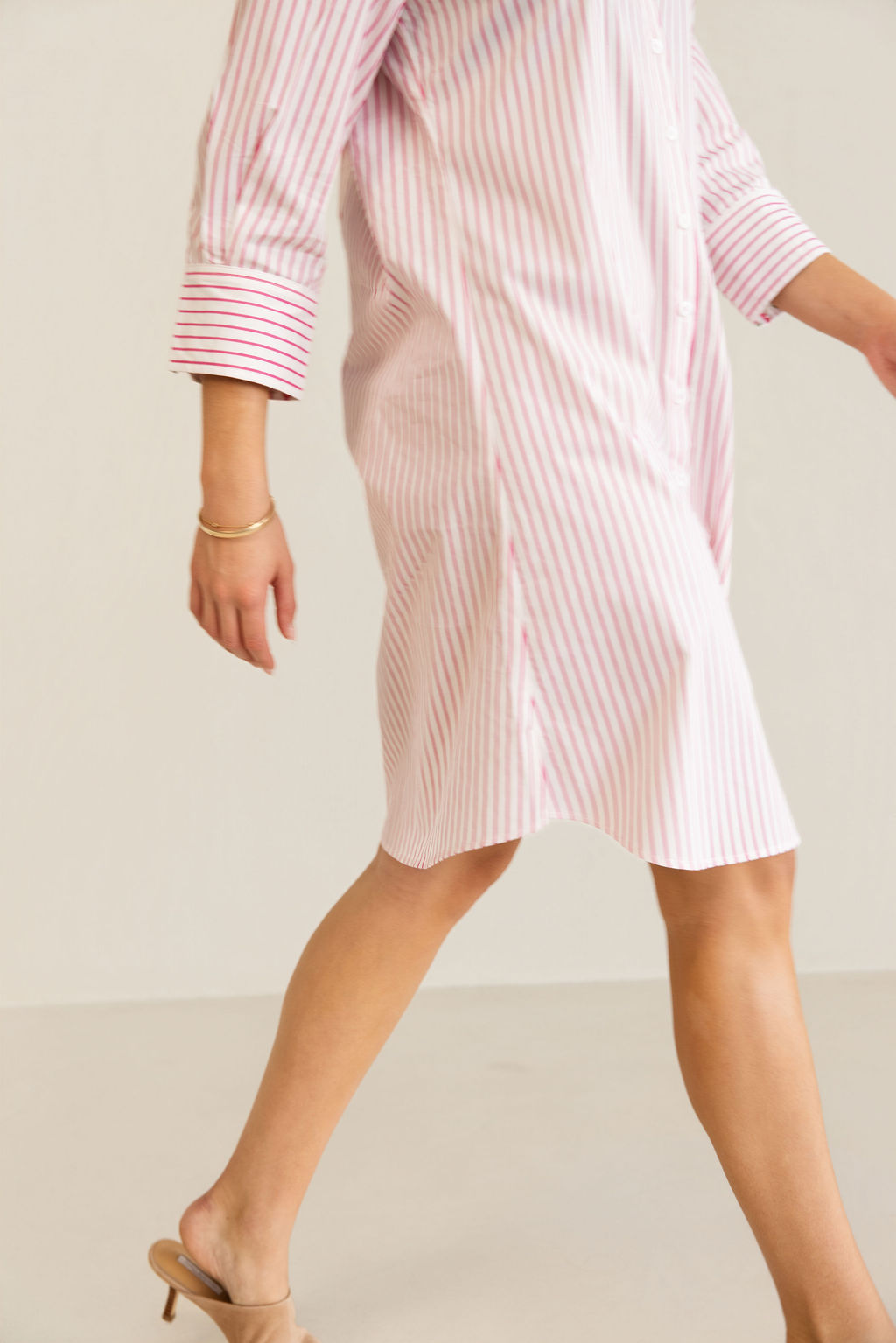 A woman modeling a pink and white Kaleido cotton vertically striped button-down shirt dress with three-quarter sleeves, standing against a plain off-white studio background.