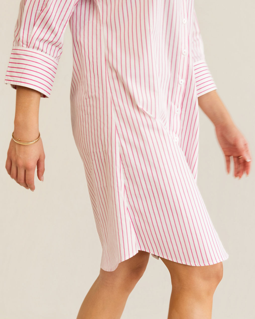 A woman modeling a pink and white Kaleido cotton vertically striped button-down shirt dress with three-quarter sleeves, standing against a plain off-white studio background.