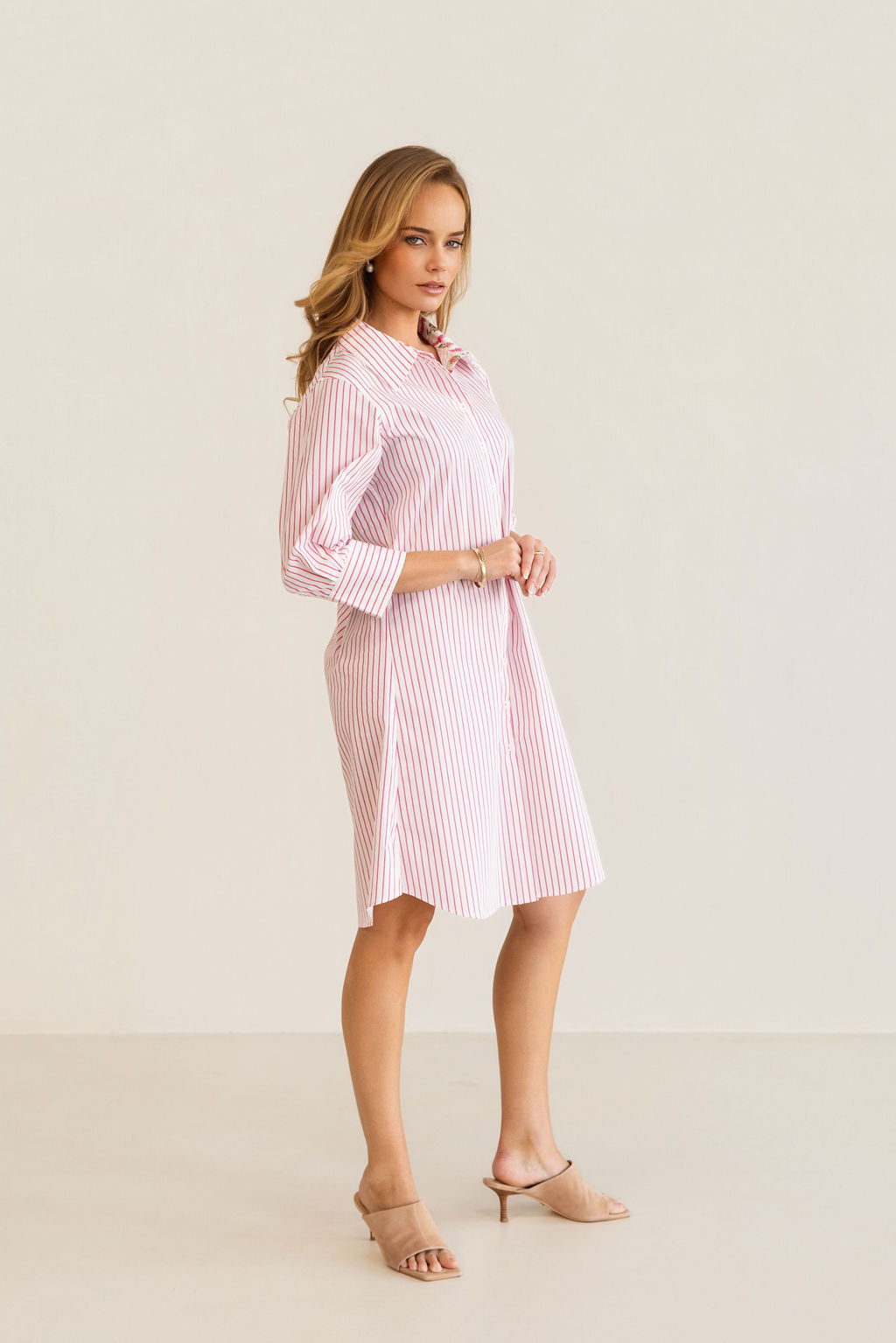 A woman modeling a pink and white Kaleido cotton vertically striped button-down shirt dress with three-quarter sleeves, standing against a plain off-white studio background.