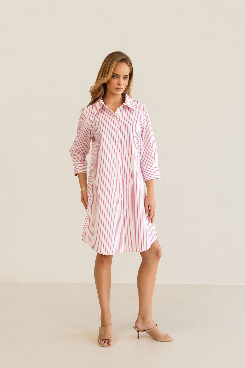 A woman modeling a pink and white Kaleido cotton vertically striped button-down shirt dress with three-quarter sleeves, standing against a plain off-white studio background.