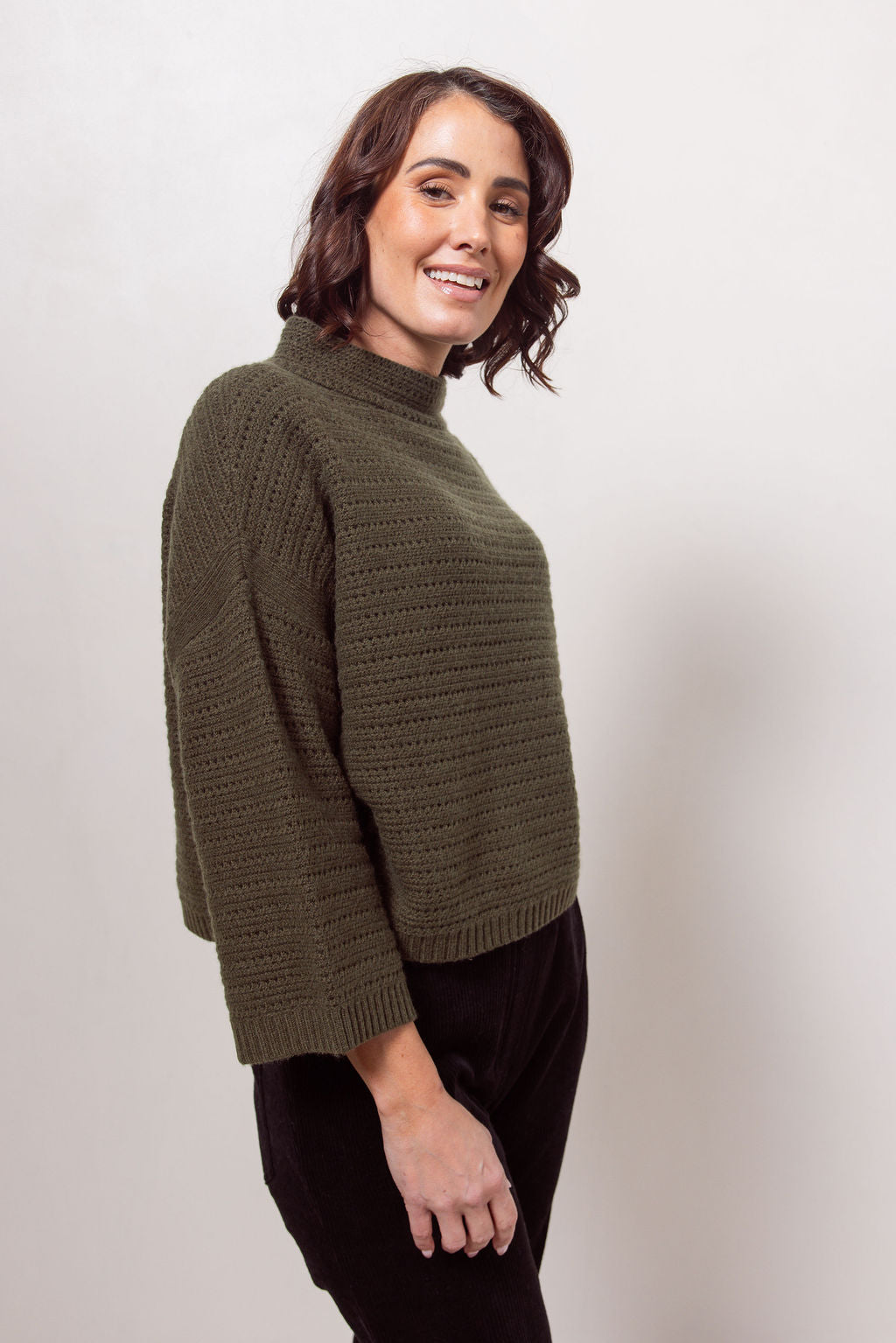 Woman wearing a See Saw Khaki pointelle sweater and black pants standing against a white background.