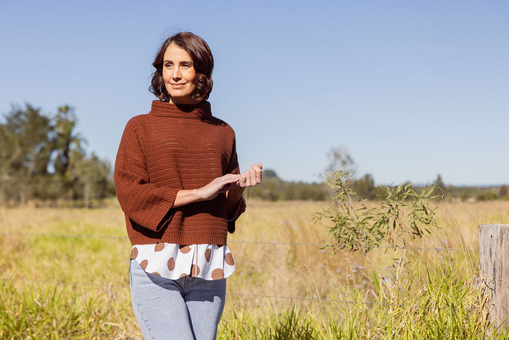 Woman wearing a See Saw copper pointelle sweater and polka dot shirt standing in a field with a clear blue sky.