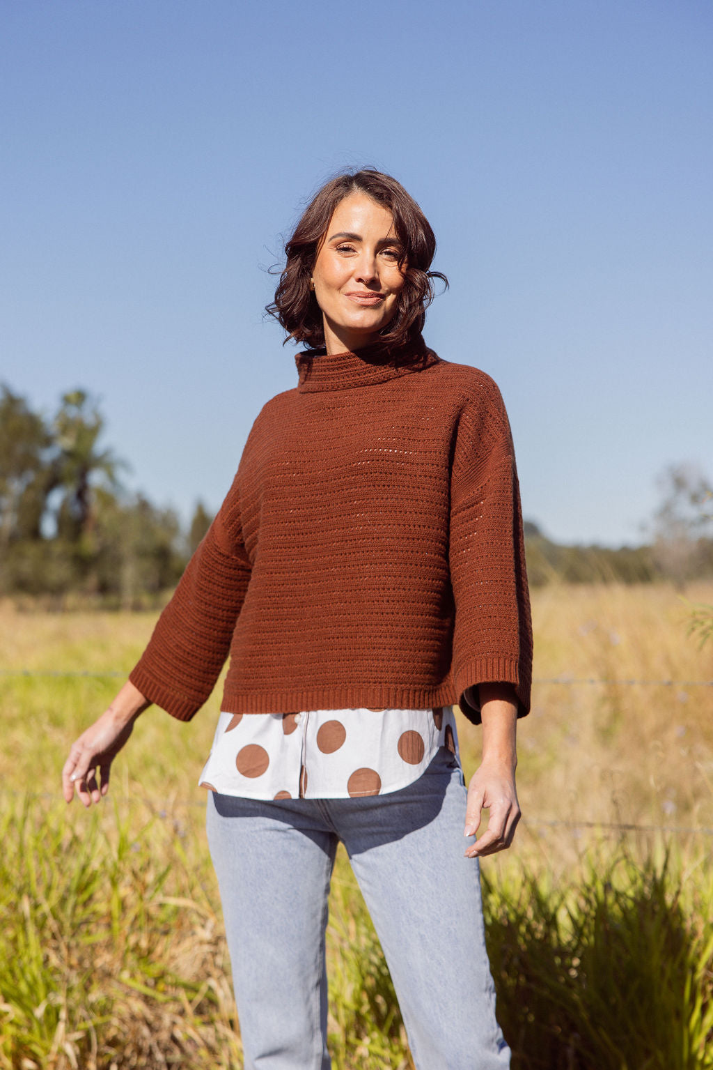Woman wearing a See Saw copper pointelle sweater and polka dot shirt standing in a field with a clear blue sky.