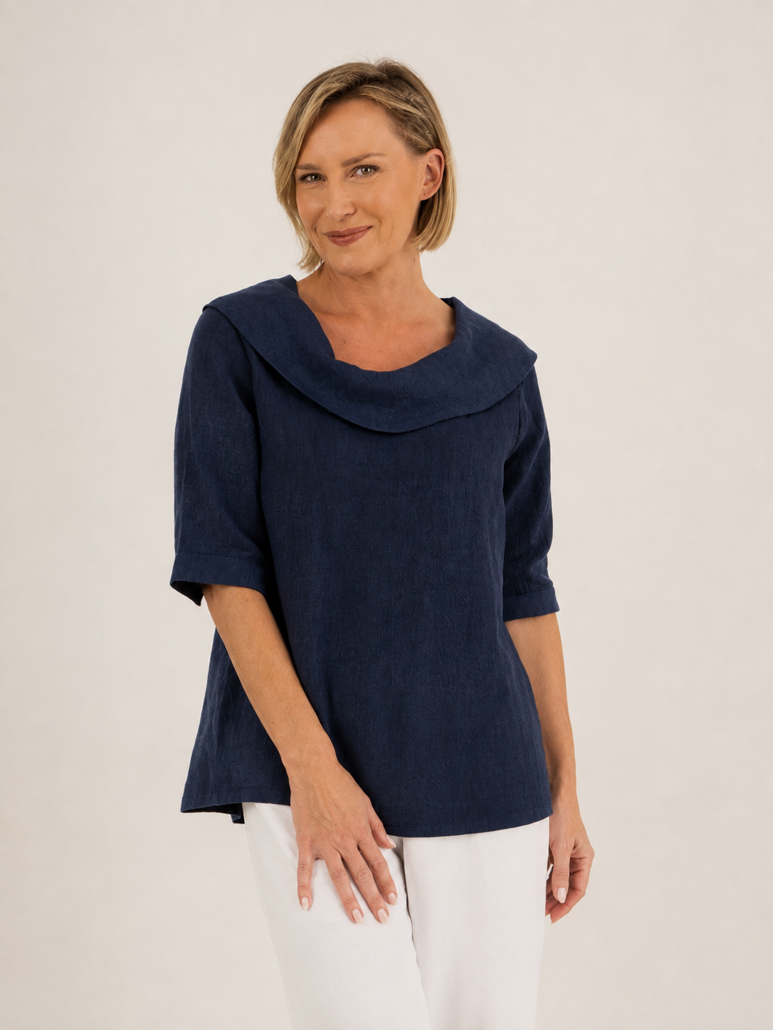 A woman modeling a navy linen See Saw top with a wide draped collar and elbow-length sleeves, paired with white pants, against a plain off-white background.