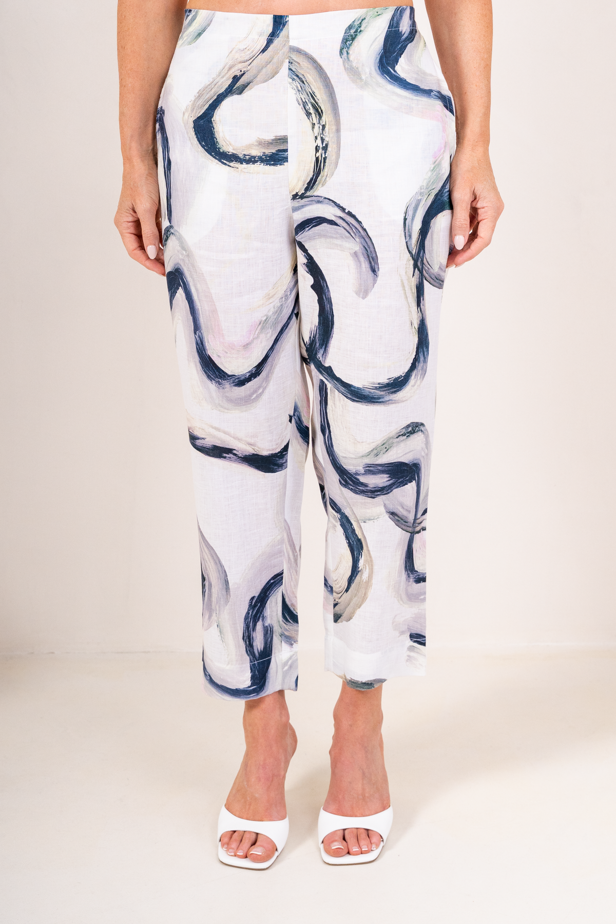 A model is shown from the waist down wearing white See Saw Flat front Riverstone Linen trousers, paired with white heeled sandals.