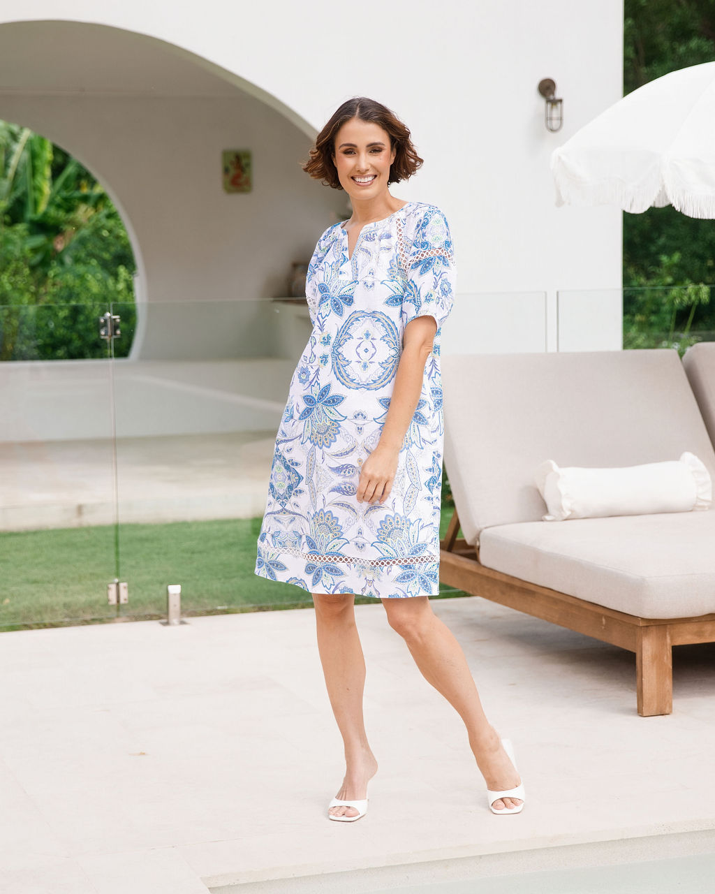 Woman modelling see saw linen dress in botanica print with lace trim in a n outdoor pool area.