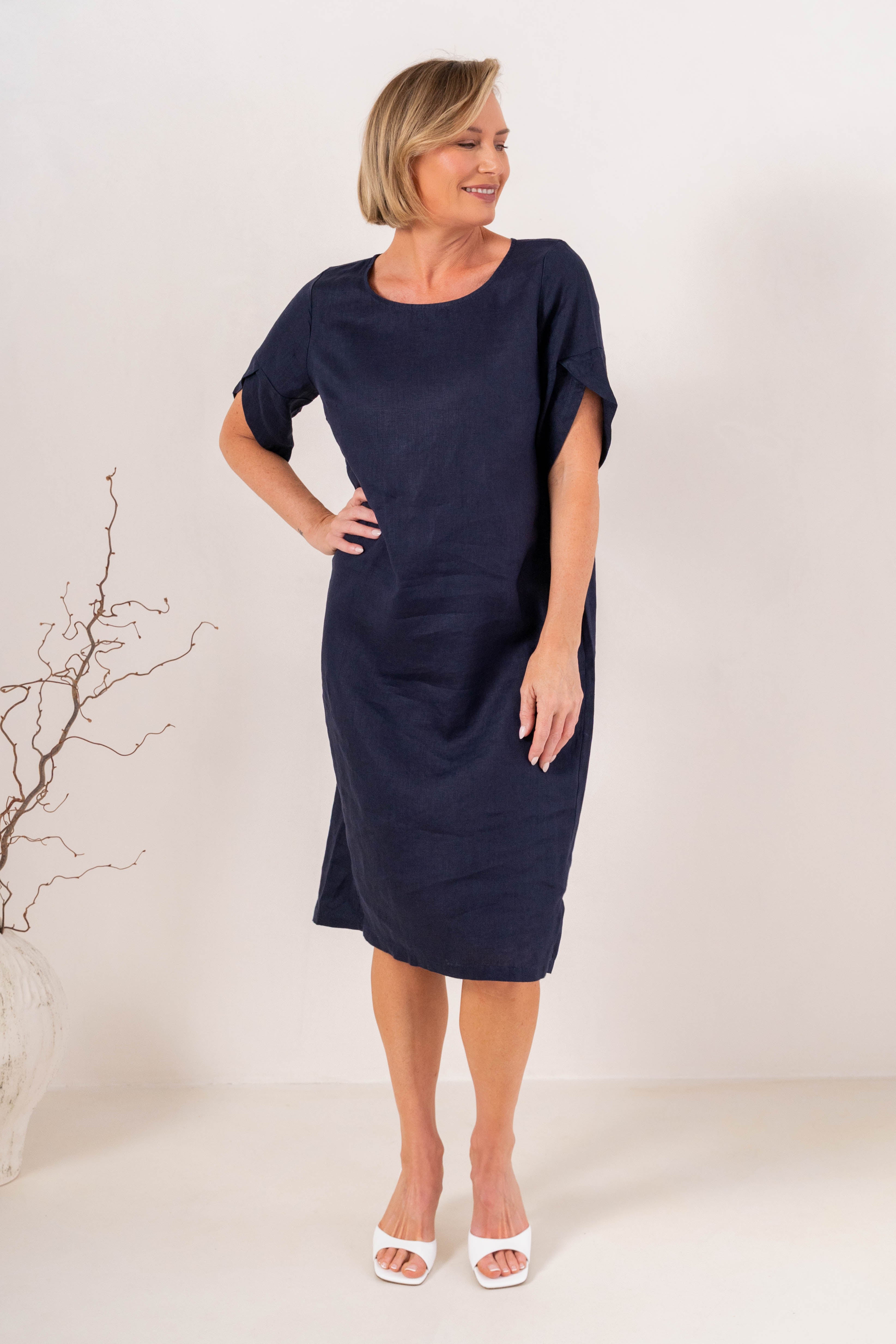 A woman modeling a See Saw Linen navy blue short-sleeve midi dress with a relaxed fit, standing against a plain off-white studio background.