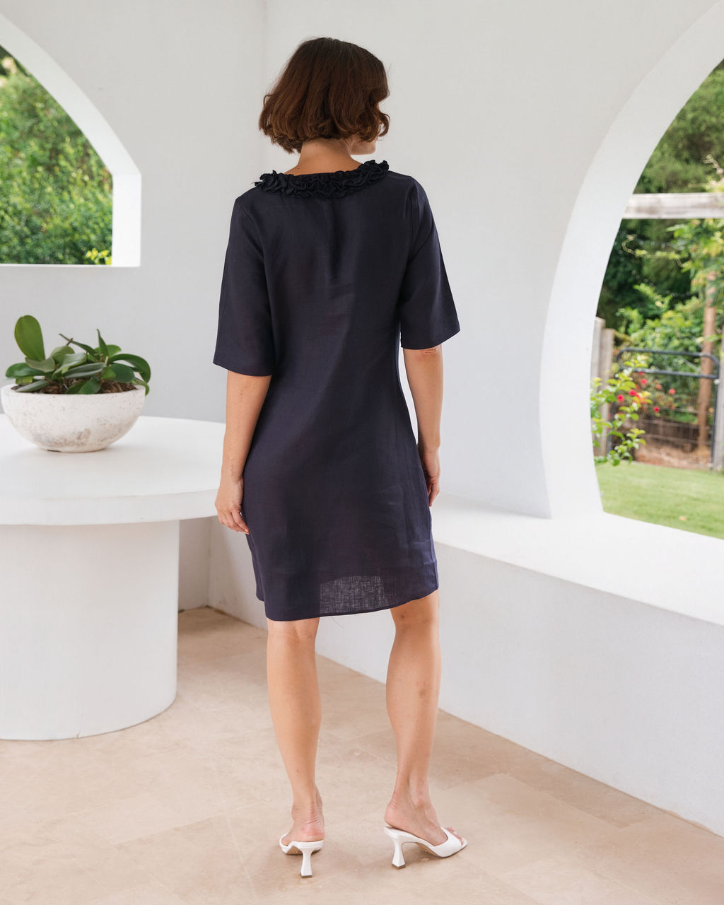 A woman wearing a dark navy blue linen See Saw dress with ruffled trim at the collar, shown from behind in a bright white architectural setting with arched windows.