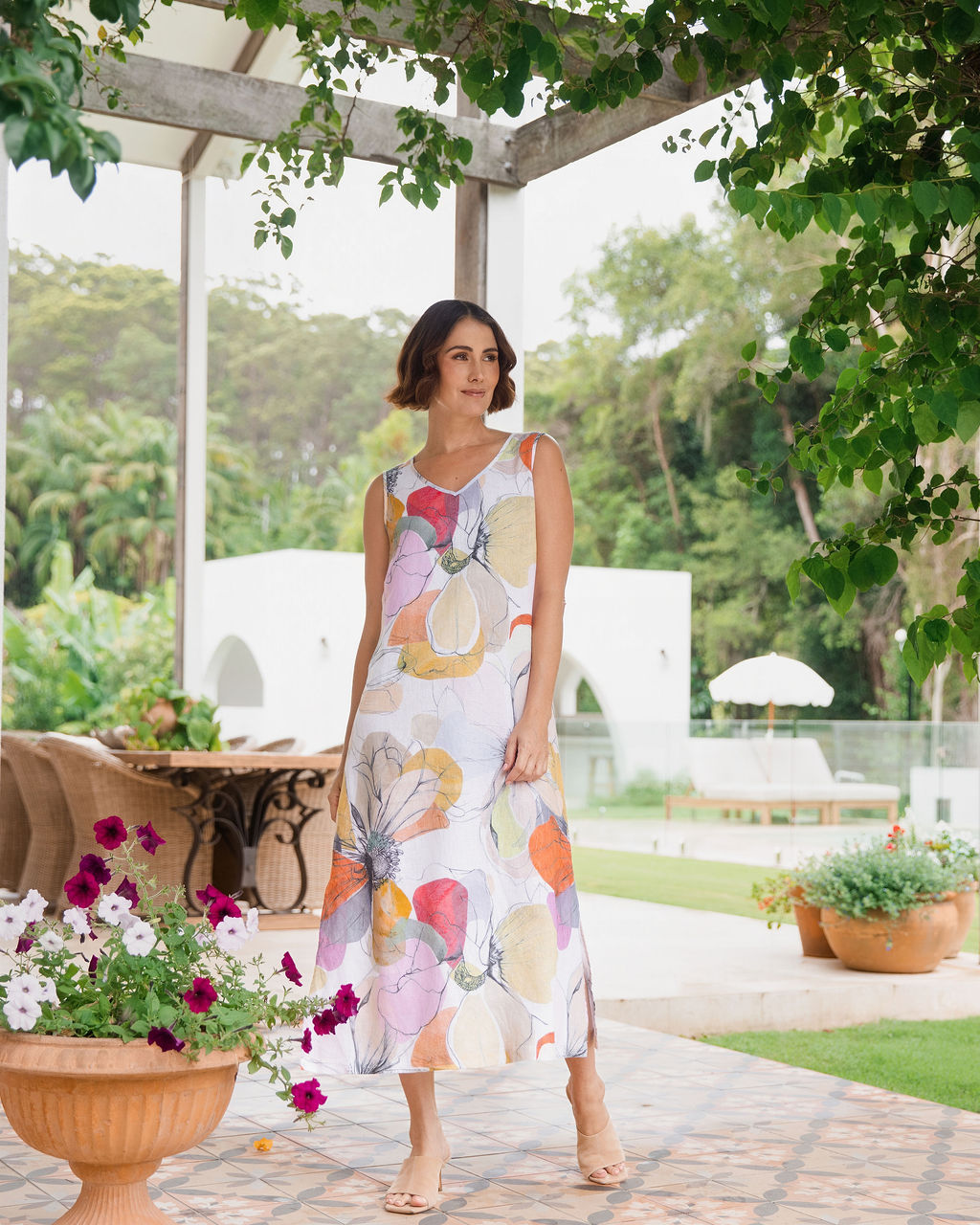 A woman wearing a sleeveless, linen See Saw knee-length A-line dress with a Petal Drift Print, standing on a tiled patio in a garden setting.