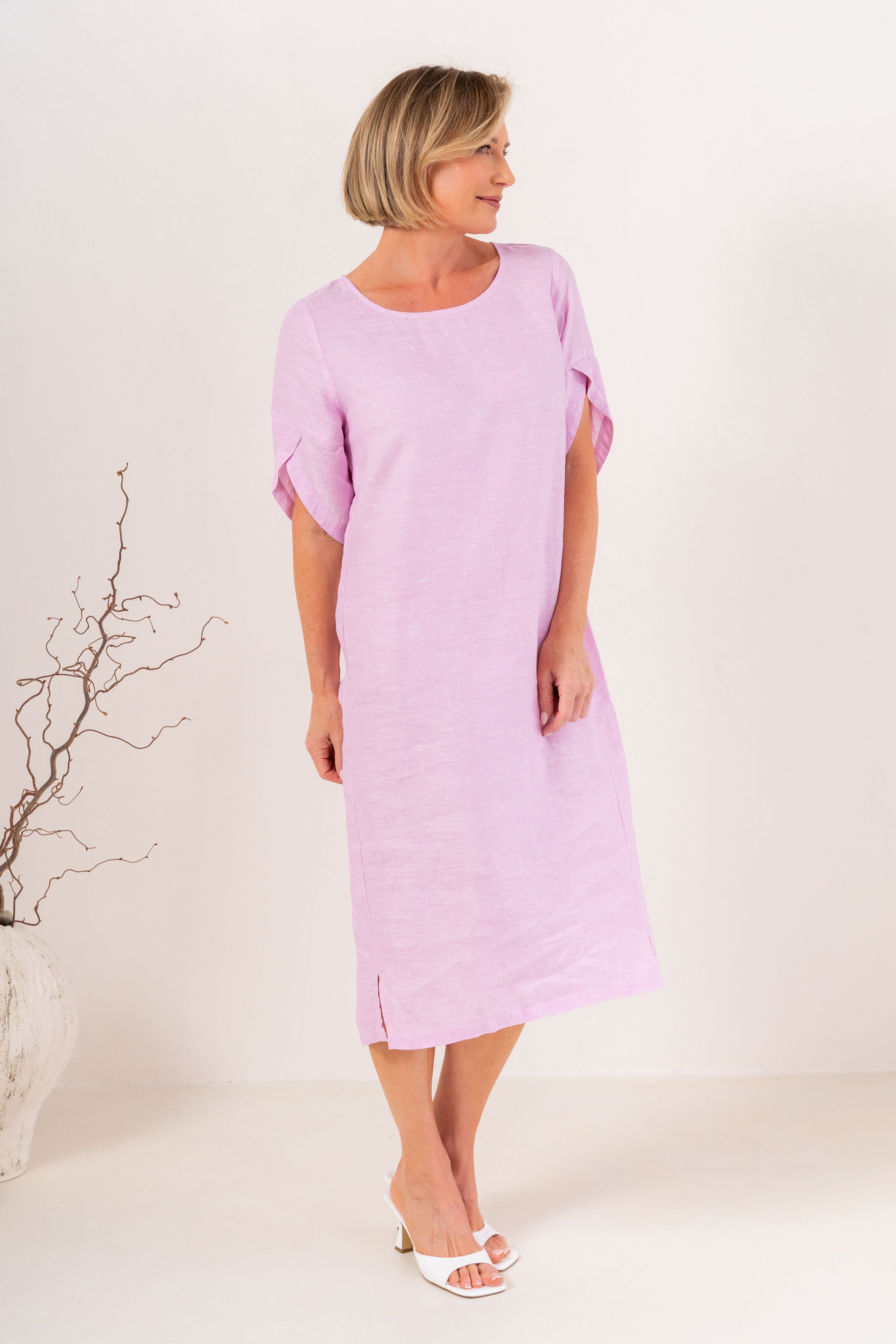 A woman modeling a See Saw Linen pink short-sleeve midi dress with a relaxed fit, standing against a plain off-white studio background.