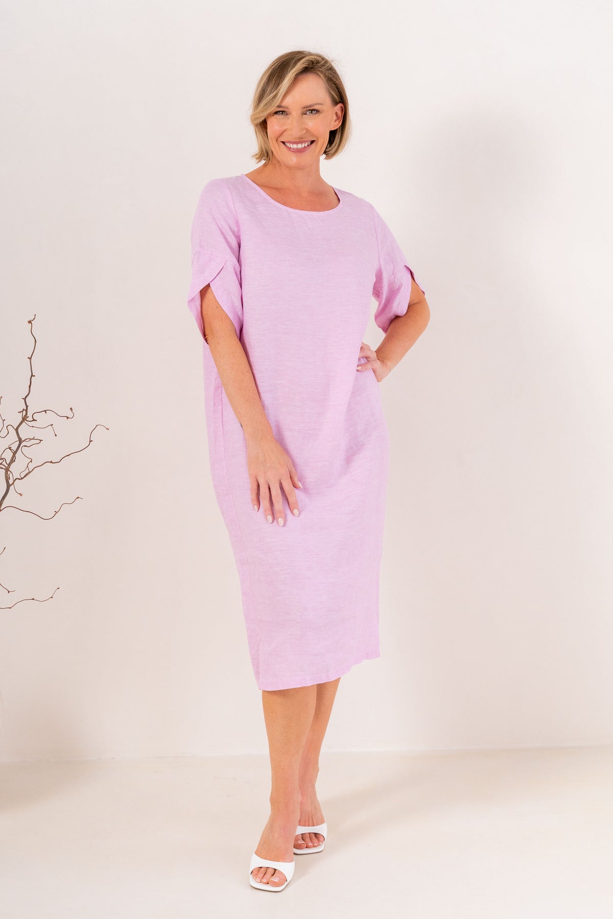 A woman modeling a See Saw Linen pink short-sleeve midi dress with a relaxed fit, standing against a plain off-white studio background.