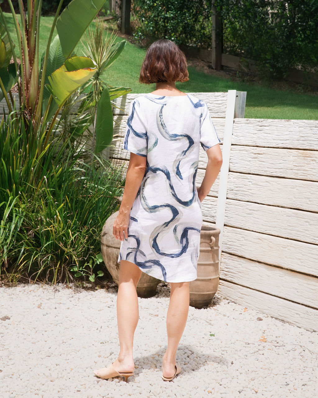 A woman stands outdoors in front of a light-colored wooden fence and greenery, wearing a See Saw short-sleeve white linen dress with a navy blue riverstone wavy pattern and pockets.