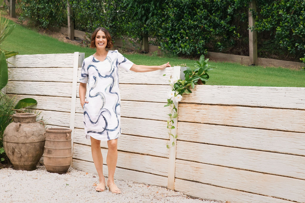 A woman stands outdoors in front of a light-colored wooden fence and greenery, wearing a See Saw short-sleeve white linen dress with a navy blue riverstone wavy pattern and pockets.
