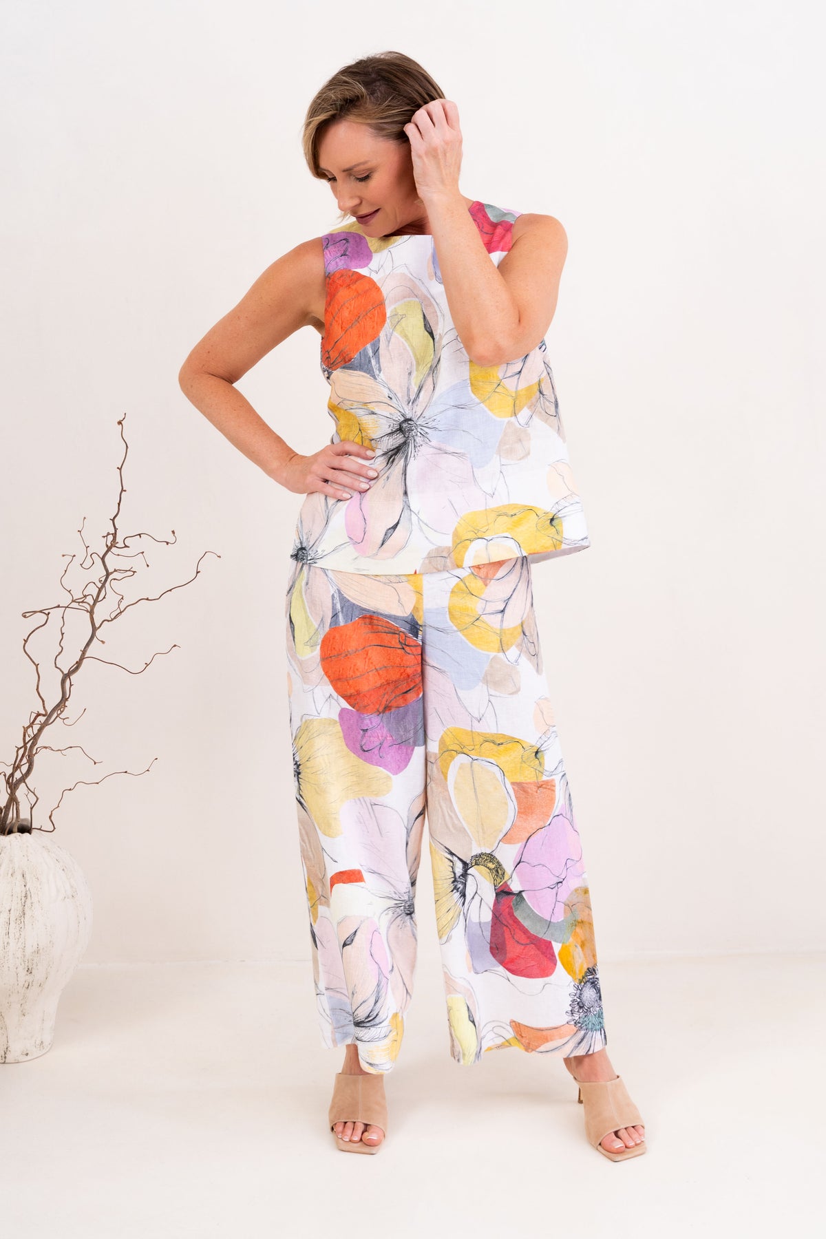 A woman modeling a sleeveless Petal Drift linen See Saw Top and pant set with a colorful abstract floral print, standing in a bright, modern living room.