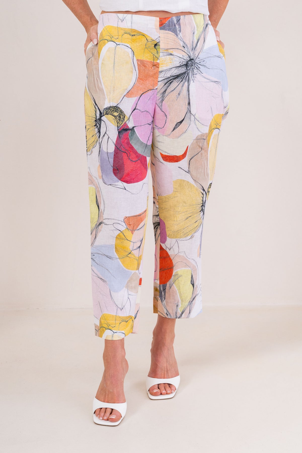 A person wearing See Saw linen cropped pants with a large abstract Petal Drift print in yellow, pink, orange, purple, and blue on a light base, paired with white heeled sandals against a plain beige studio background.