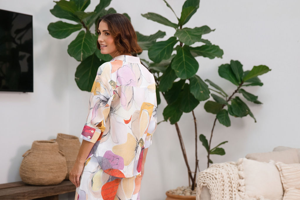 A woman modeling a matching two-piece See Saw linen set with petal drift floral print, standing in a cozy, modern living room setting with indoor plants and a sofa.