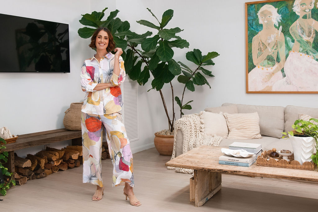A woman modeling a matching two-piece See Saw linen set with petal drift floral print, standing in a cozy, modern living room setting with indoor plants and a sofa.