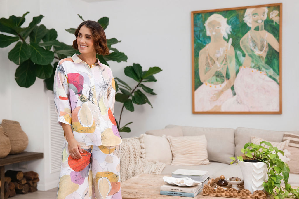 A woman modeling a matching two-piece See Saw linen set with petal drift floral print, standing in a cozy, modern living room setting with indoor plants and a sofa.