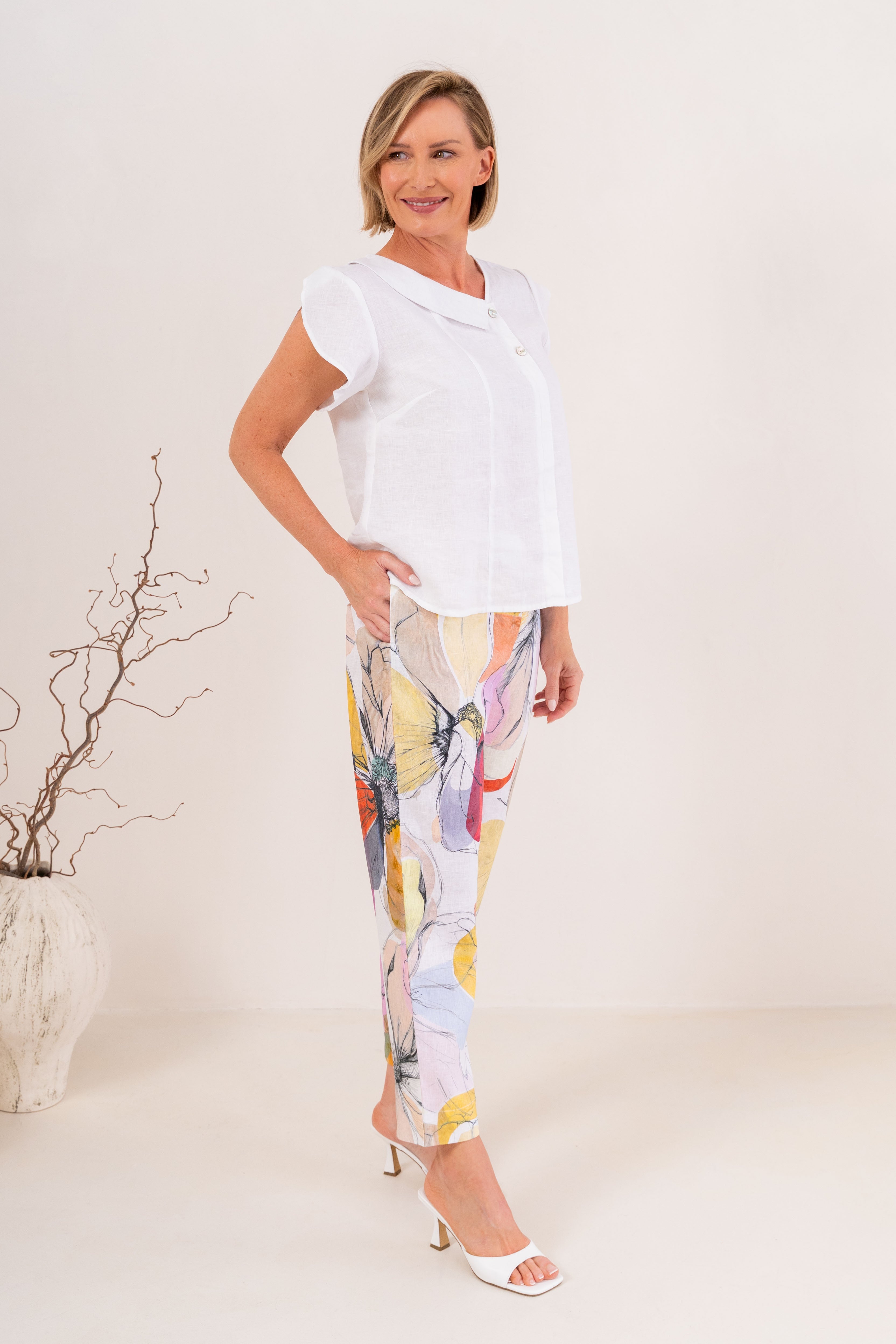 A woman modeling a white short-sleeve button-up top and high-waisted cropped trousers with a colorful abstract floral print, standing against a plain off-white studio background.