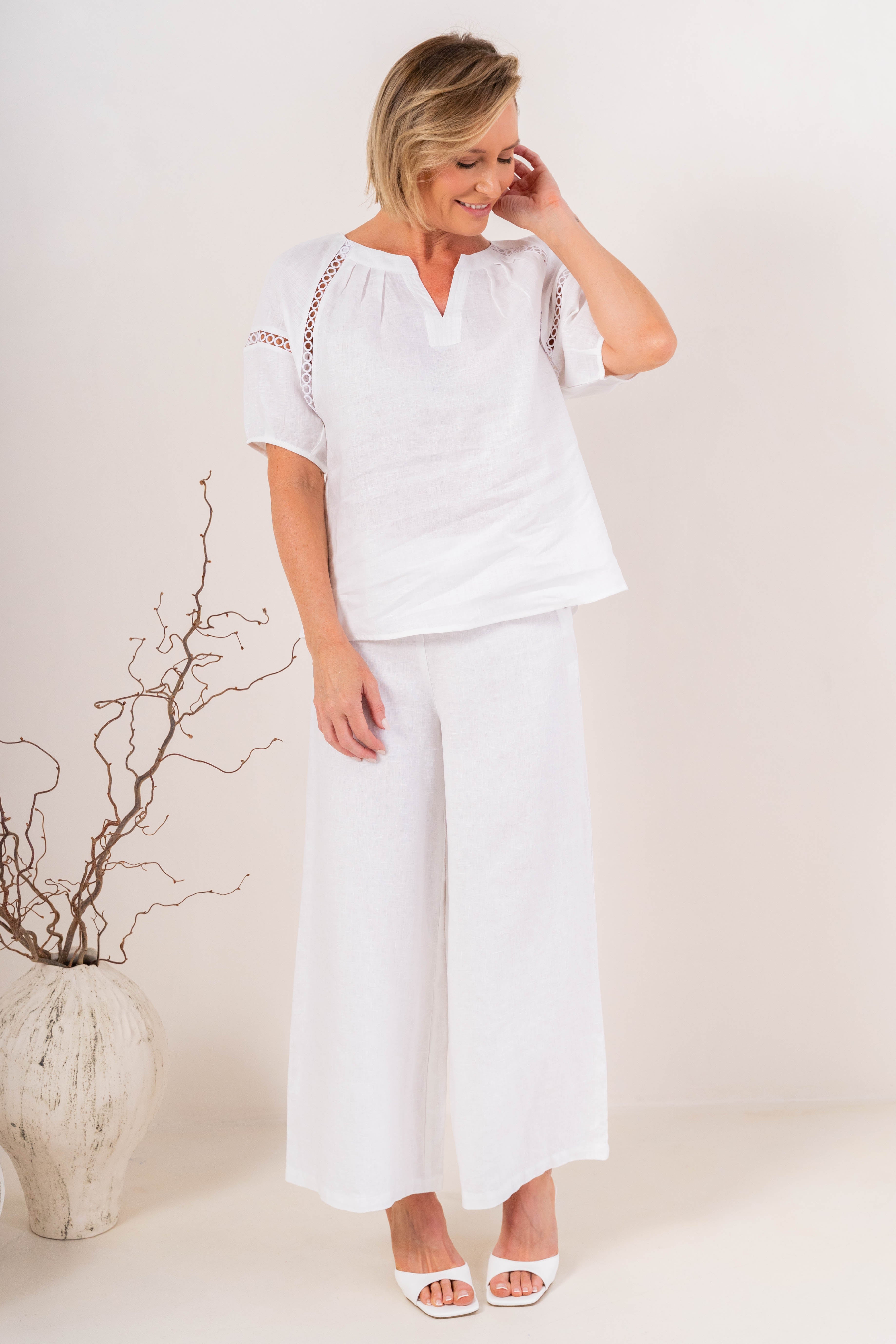 A woman modeling a white short-sleeve linen See Saw top with a split V-neck and decorative openwork lace trim along the shoulders, paired with white pants against a plain light background.