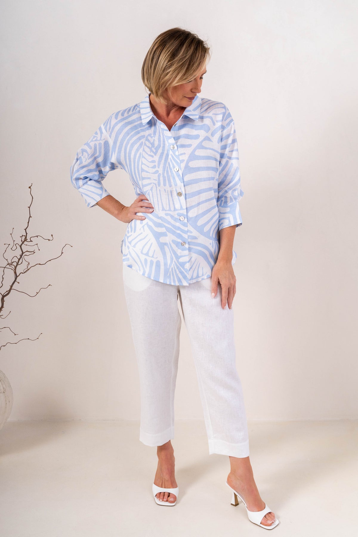 A woman modeling a light blue and white linen See Saw patterned button-up shirt with three-quarter sleeves, paired with white pants, against a plain light background.