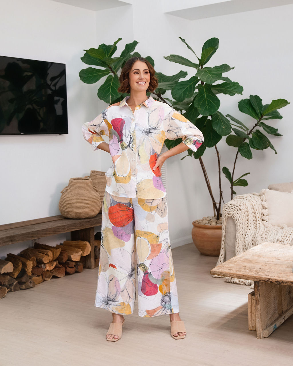 A woman modeling a matching two-piece See Saw linen set with petal drift floral print, standing in a cozy, modern living room setting with indoor plants and a sofa.