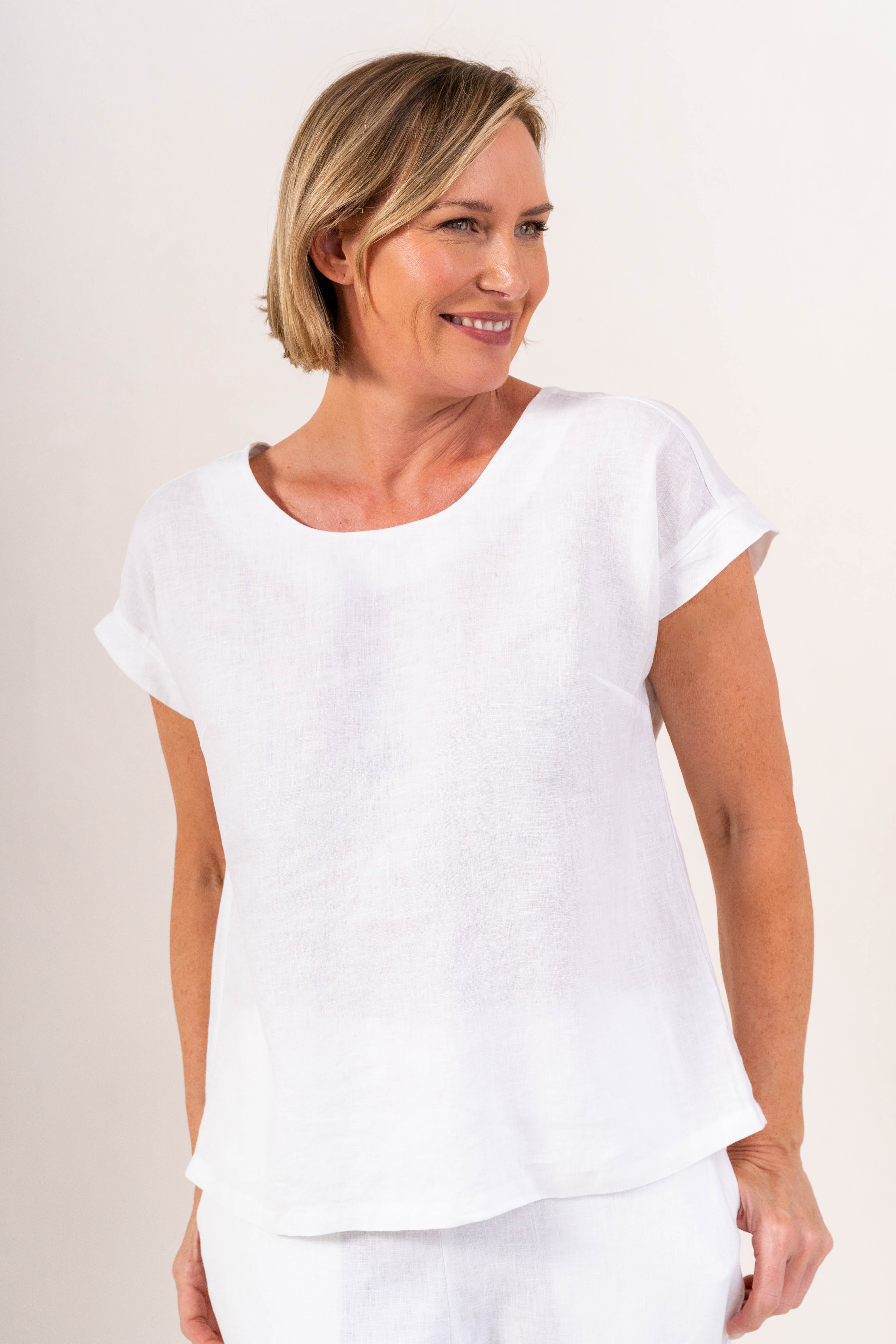 A smiling blonde woman wearing a white short-sleeve See Saw linen top and matching white pants, photographed against a plain light beige studio background.