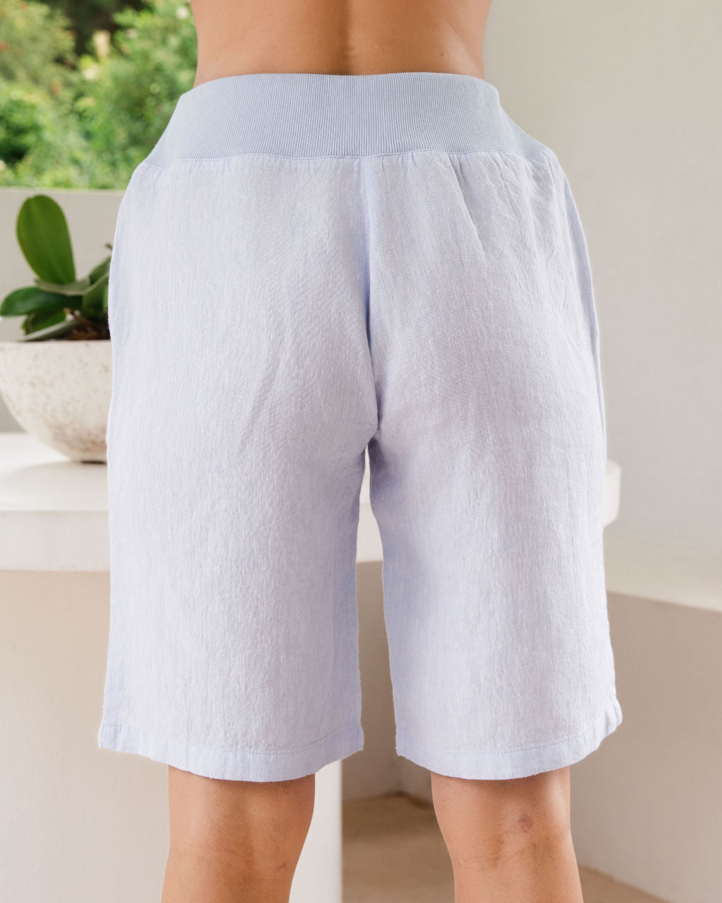 Light blue linen-blend See Saw linen shorts with a contrasting light blue elastic waistband, shown on a model from the waist down in a bright indoor setting.