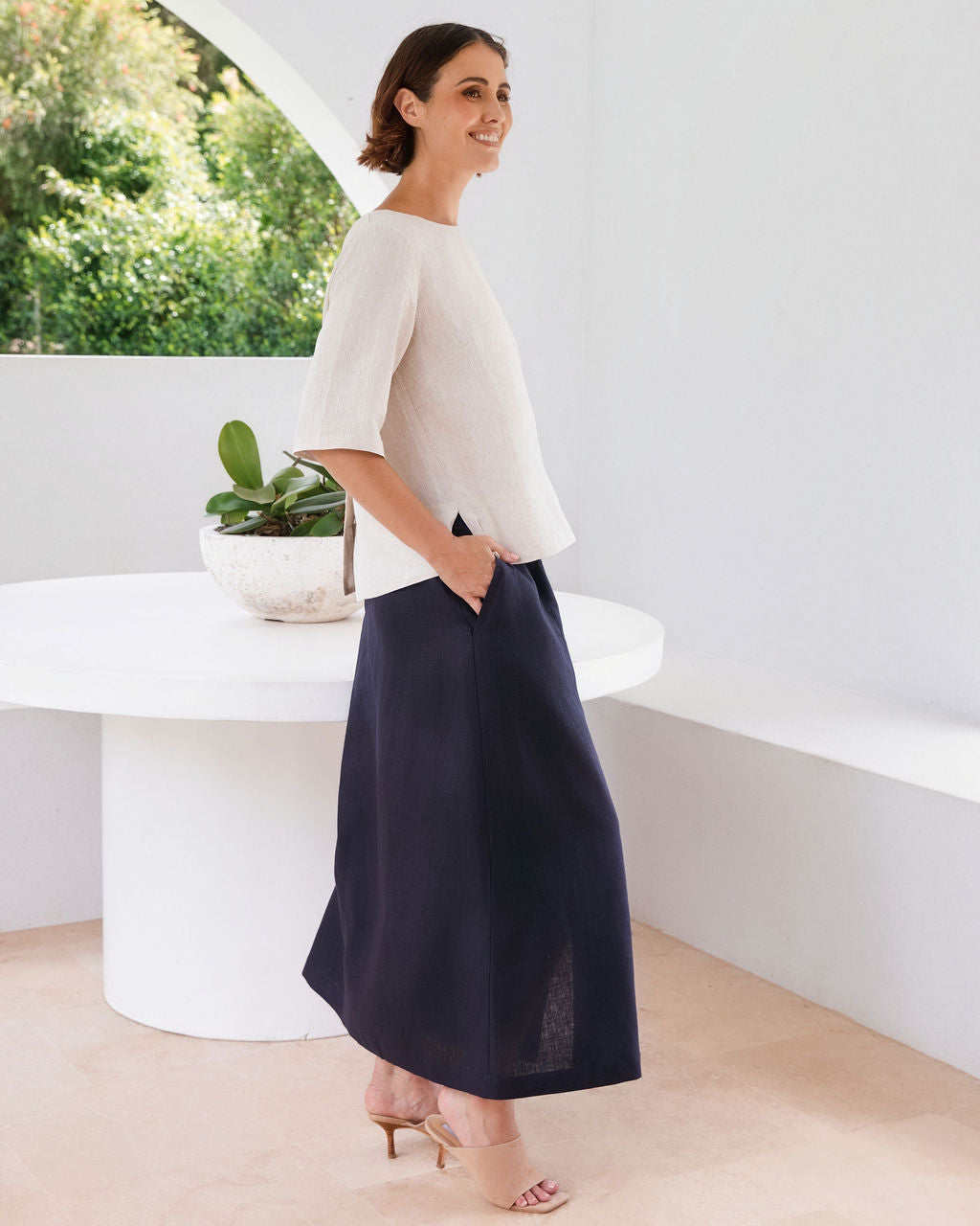 A woman modeling a cream short-sleeve linen top and a navy blue midi skirt, standing in a bright, modern white room with a curved white table and a potted plant in the background.