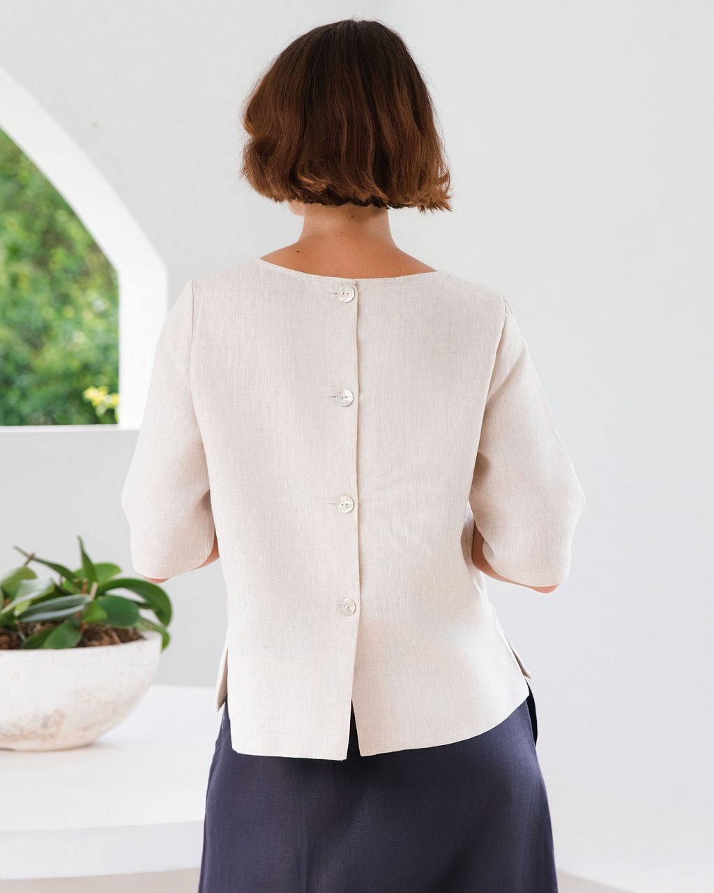 A woman modeling a natural linen See Saw three-quarter sleeve top and light white skirts , standing in a bright, modern white room with a round white table.