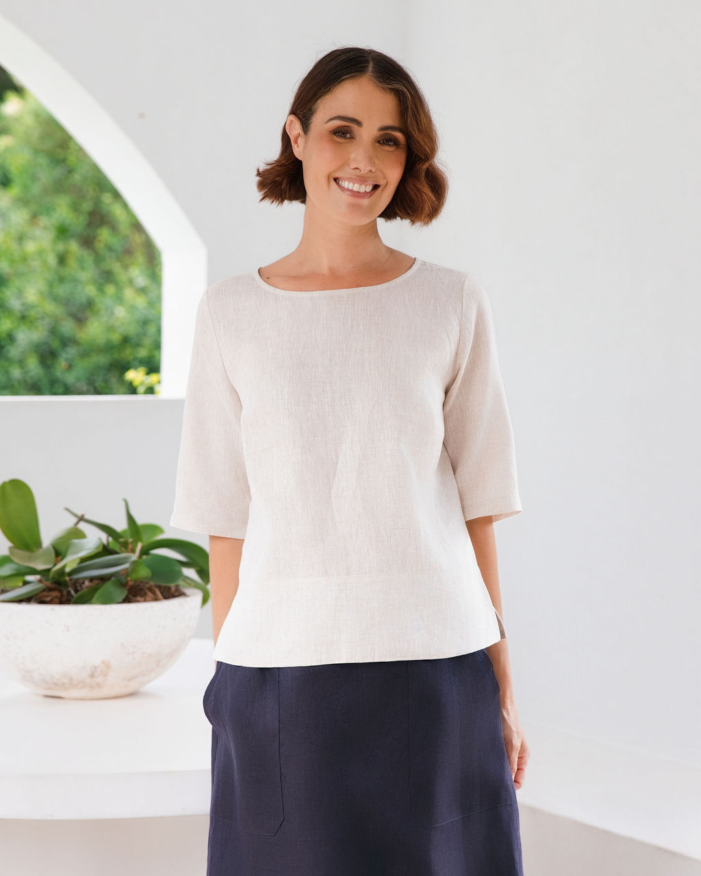 A woman wearing a See Saw cream-colored linen top with elbow-length sleeves and a navy blue skirt, standing in a bright, modern white room with a potted plant in the background.