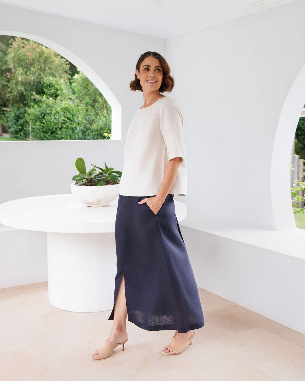 A woman modeling a natural linen See Saw three-quarter sleeve top and light white skirts , standing in a bright, modern white room with a round white table.
