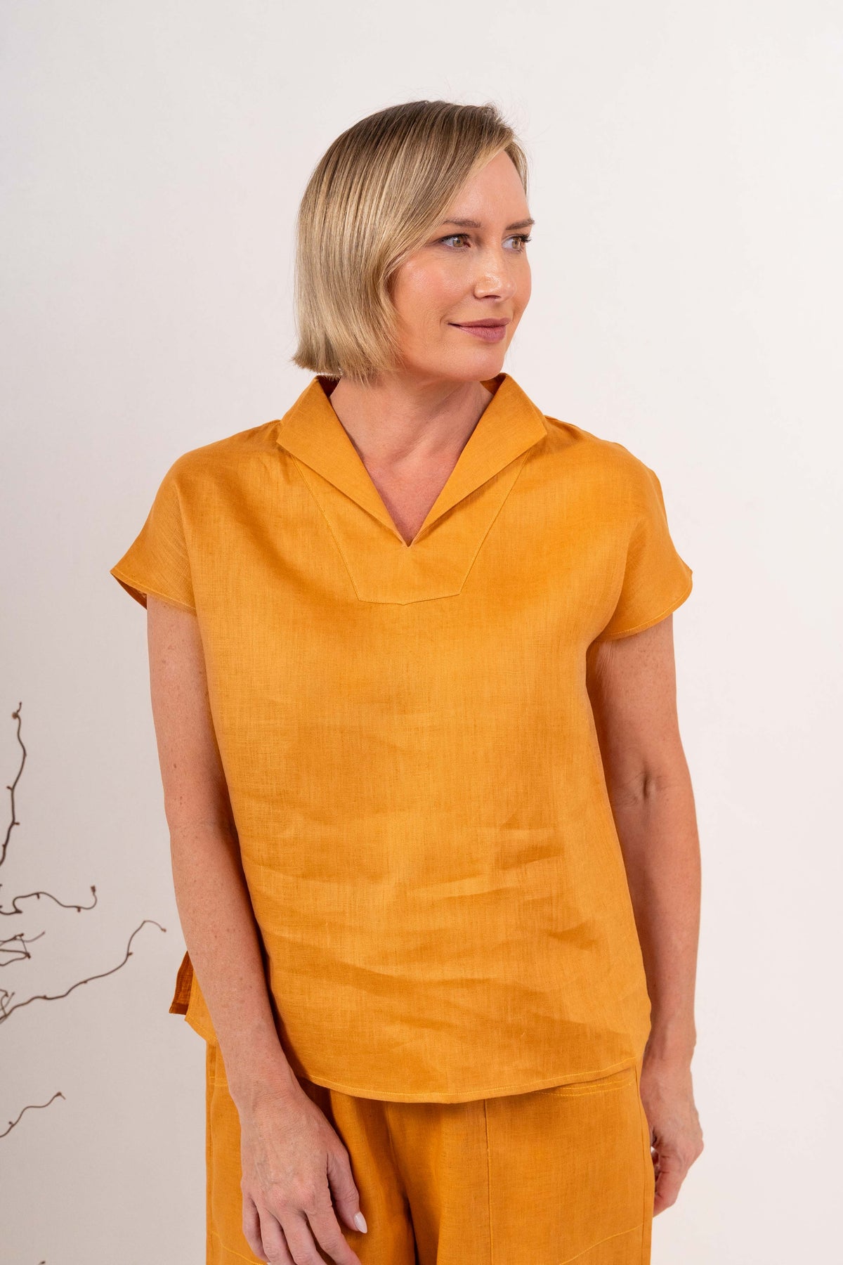 A woman modeling a marigold See Saw linen short-sleeve top with a split V-neck collar, paired with matching yellow linen pants, against a light off-white background.