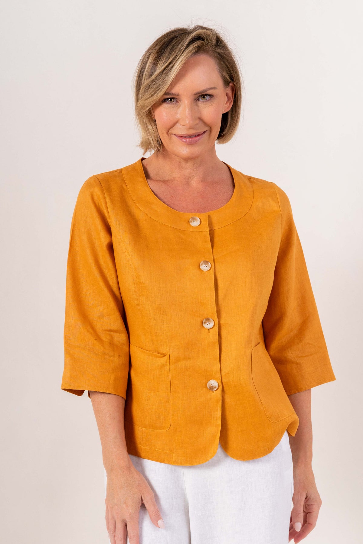 Woman modelling marigold linen see saw utility jacket top and white pants and against a plain background.