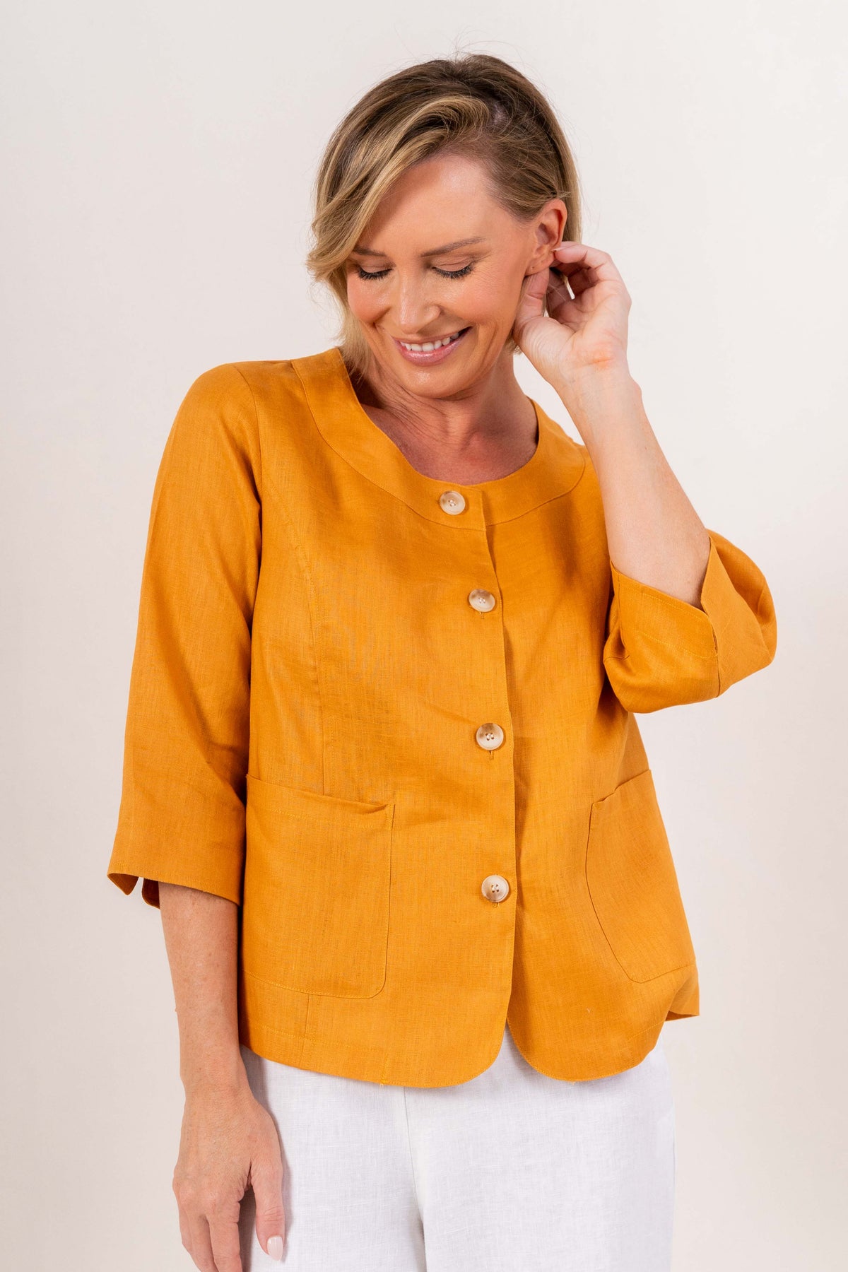 Woman modelling marigold linen see saw utility jacket top and white pants and against a plain background.