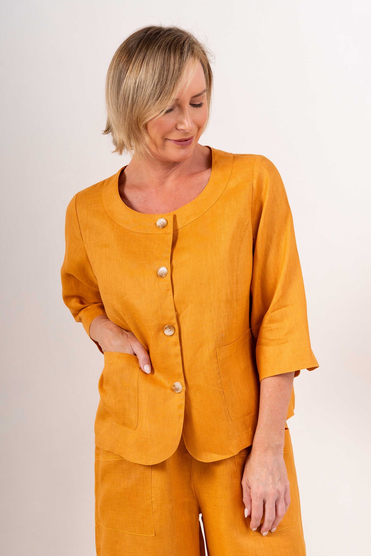 Woman modelling marigold linen see saw utility jacket top and white pants and against a plain background.