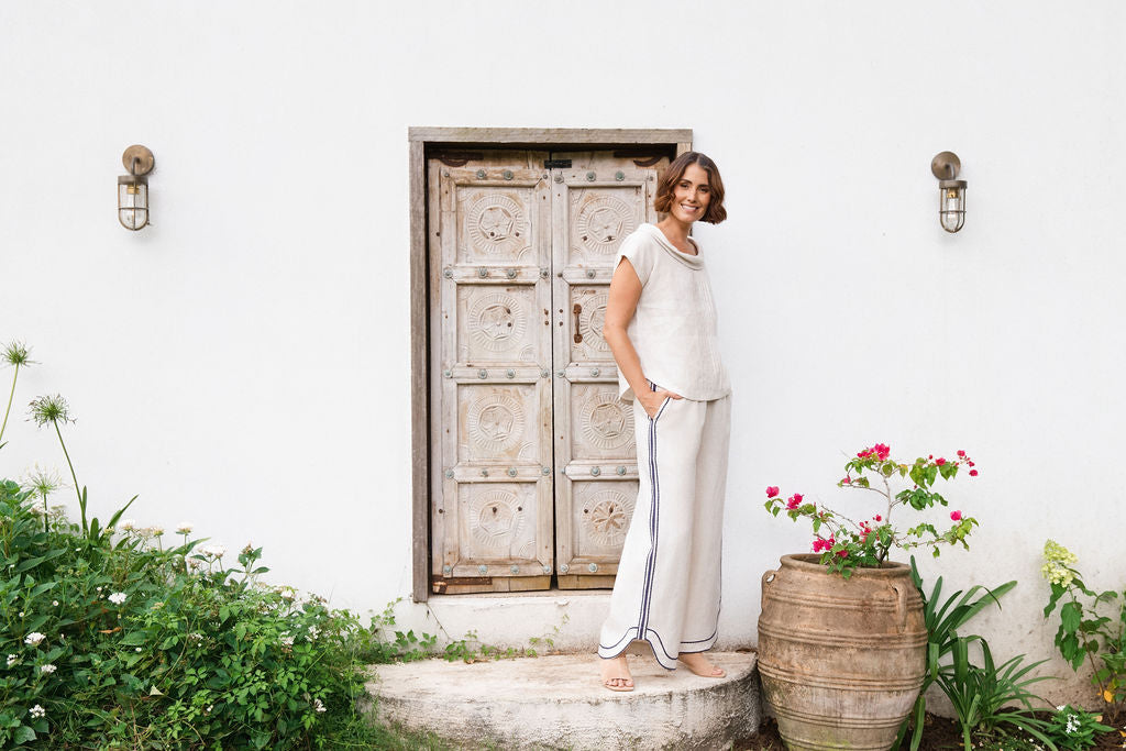 A woman stands barefoot on a stone step in front of a white wall and rustic wooden door, wearing a See Saw linen natural -colored short-sleeve knit top and matching wide-leg pants with dark piping.
