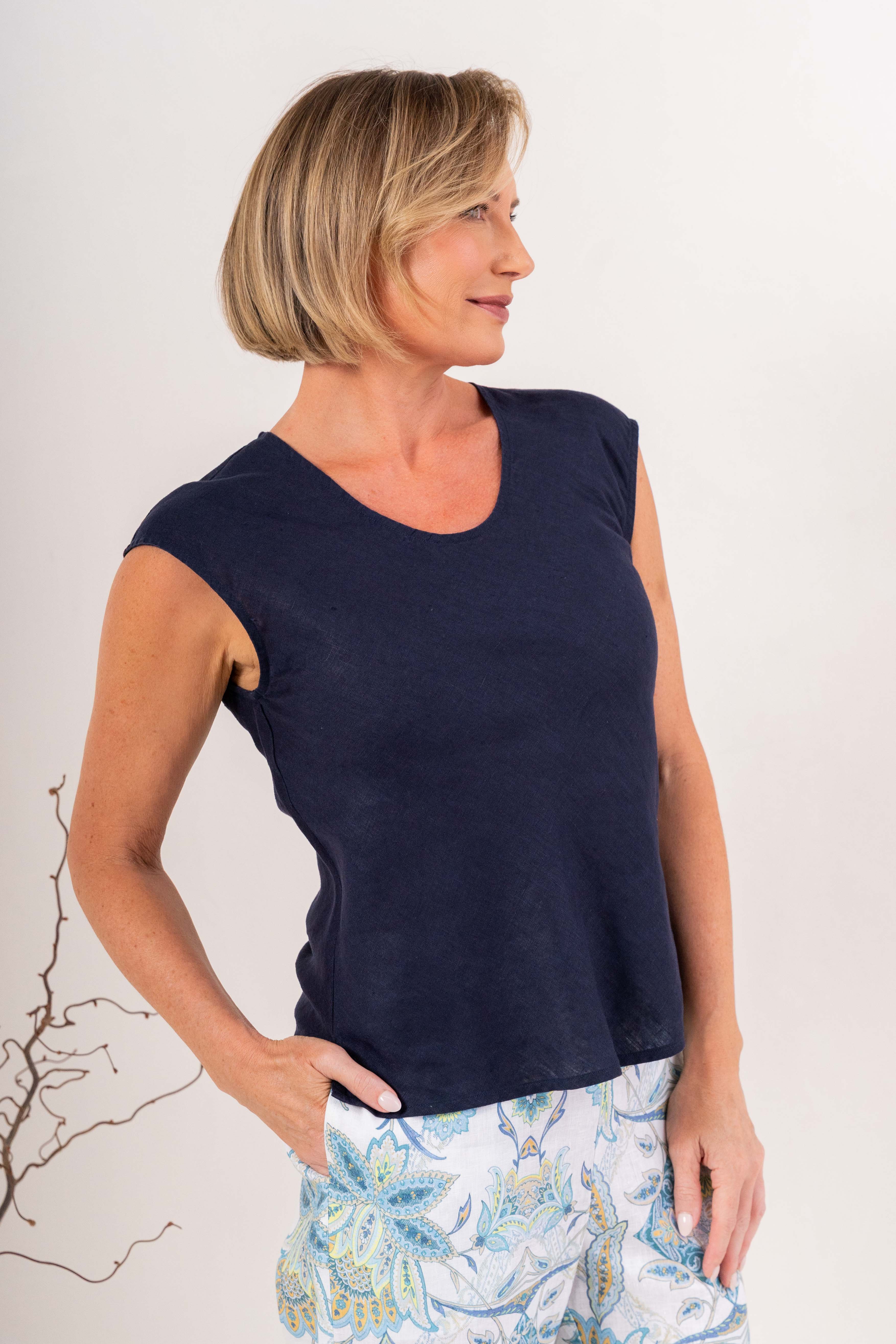 A smiling blonde woman wearing a navy sleeveless V-neck top in linen by See Saw and matching white pants, standing against a plain light background.