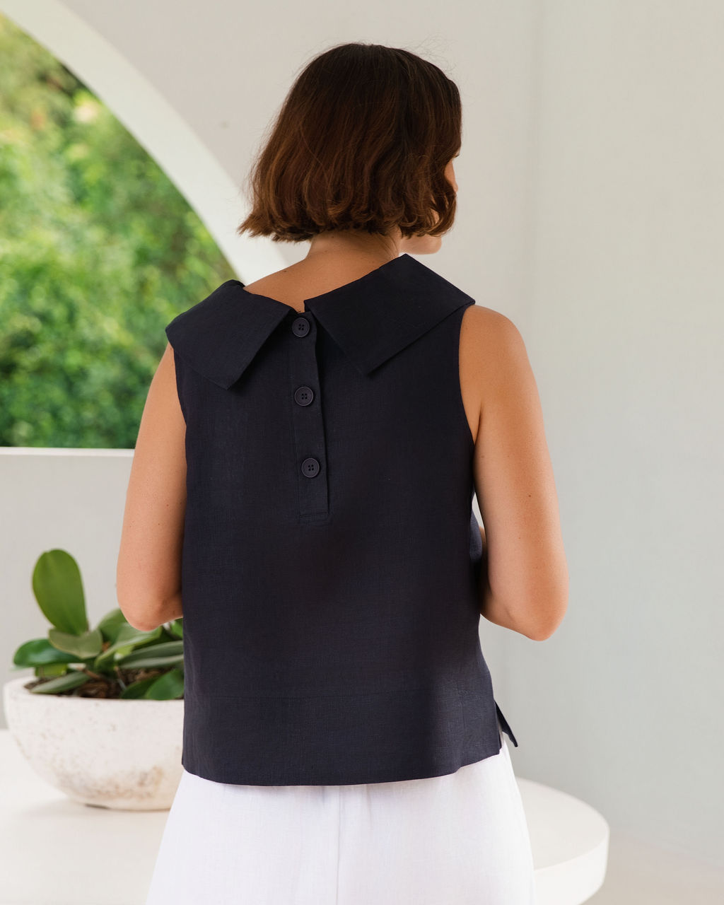 A woman viewed from behind wearing a sleeveless dark navy See Saw Linen top e with a large pointed collar and three buttons down the back, paired with white bottoms in a bright indoor setting with a plant visible.