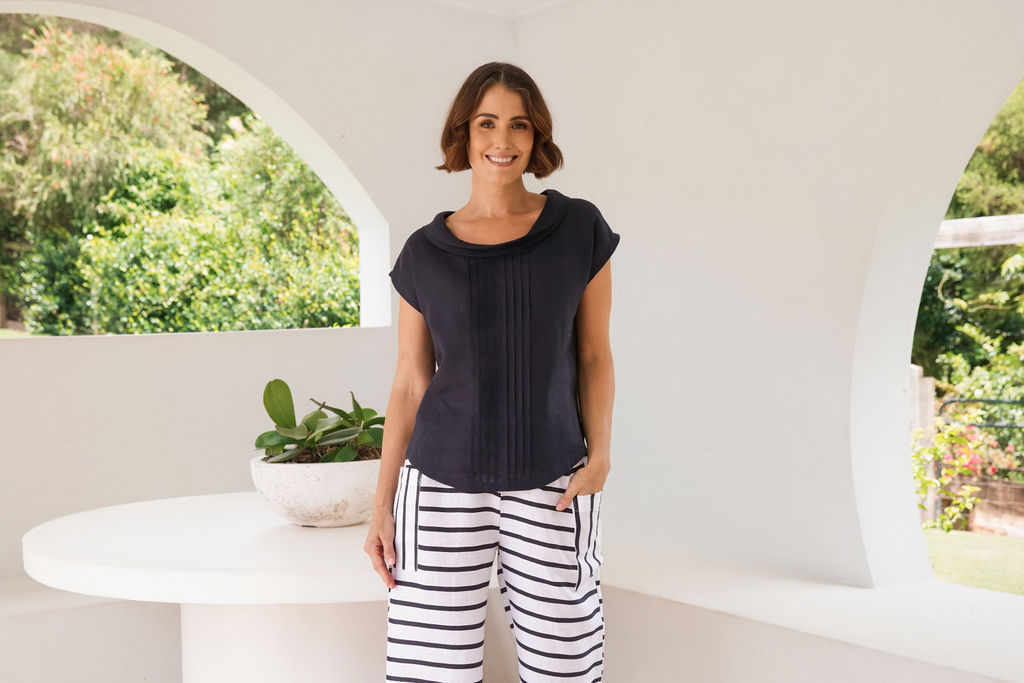 A woman modeling a navy See Saw linen sleeveless top and white stripe pants set in a bright, architectural outdoor space with a wooden coffee table, sofa, and potted plants.