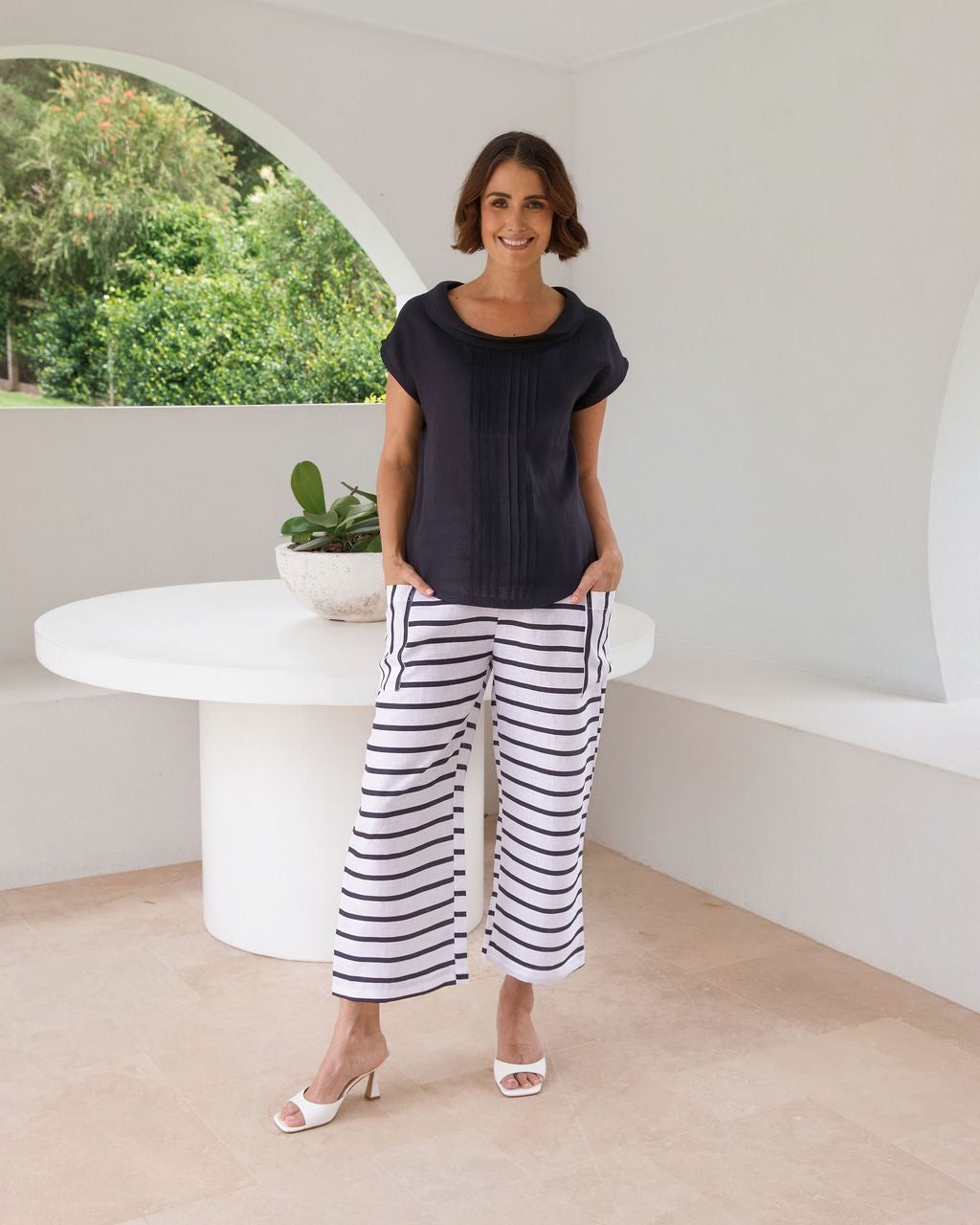 A woman stands in a bright, modern white architectural space with arched windows overlooking greenery, wearing a dark navy linen See Saw top sleeveless top and navy-and-white horizontally striped cropped pants.