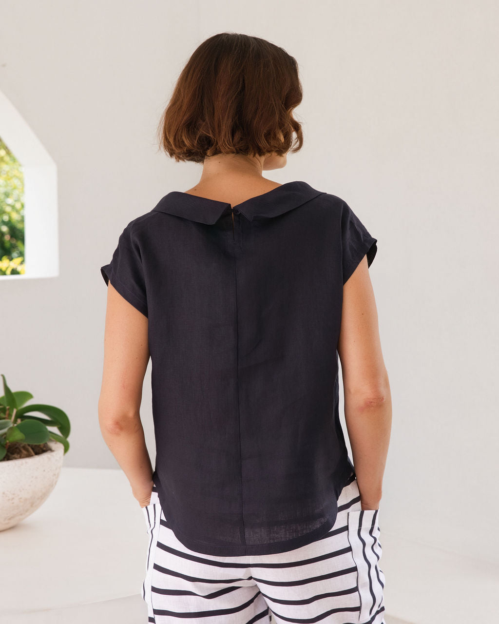 A woman stands in a bright, modern white architectural space with arched windows overlooking greenery, wearing a dark navy linen See Saw top sleeveless top and navy-and-white horizontally striped cropped pants.