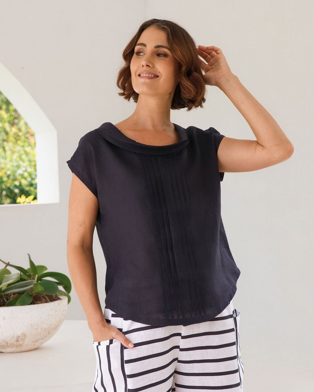 A woman stands in a bright, modern white architectural space with arched windows overlooking greenery, wearing a dark navy linen See Saw top sleeveless top and navy-and-white horizontally striped cropped pants.