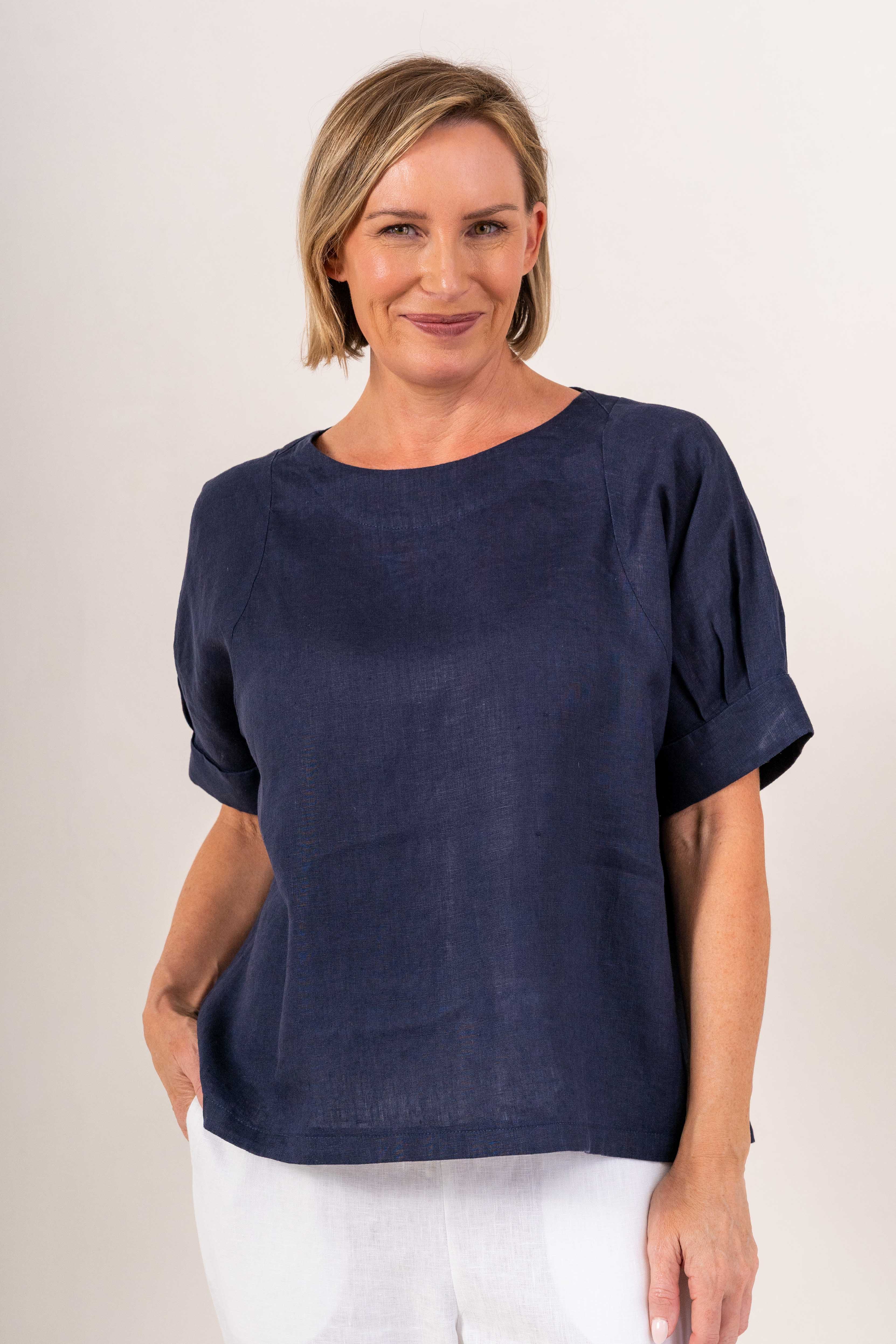 A woman wearing a See Saw Linen navy blue short-sleeve linen top with white pants, standing against a plain light beige background.