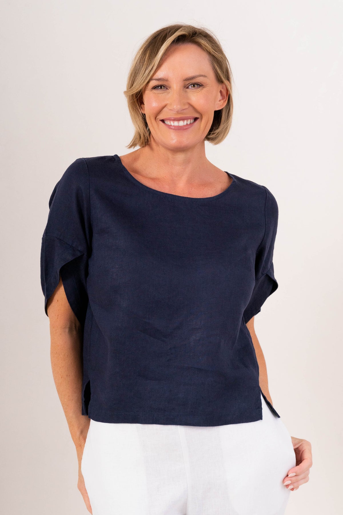 A woman wearing a navy See Saw linen short-sleeve top with ruffled or layered sleeve details and white pants, photographed against a plain light background.