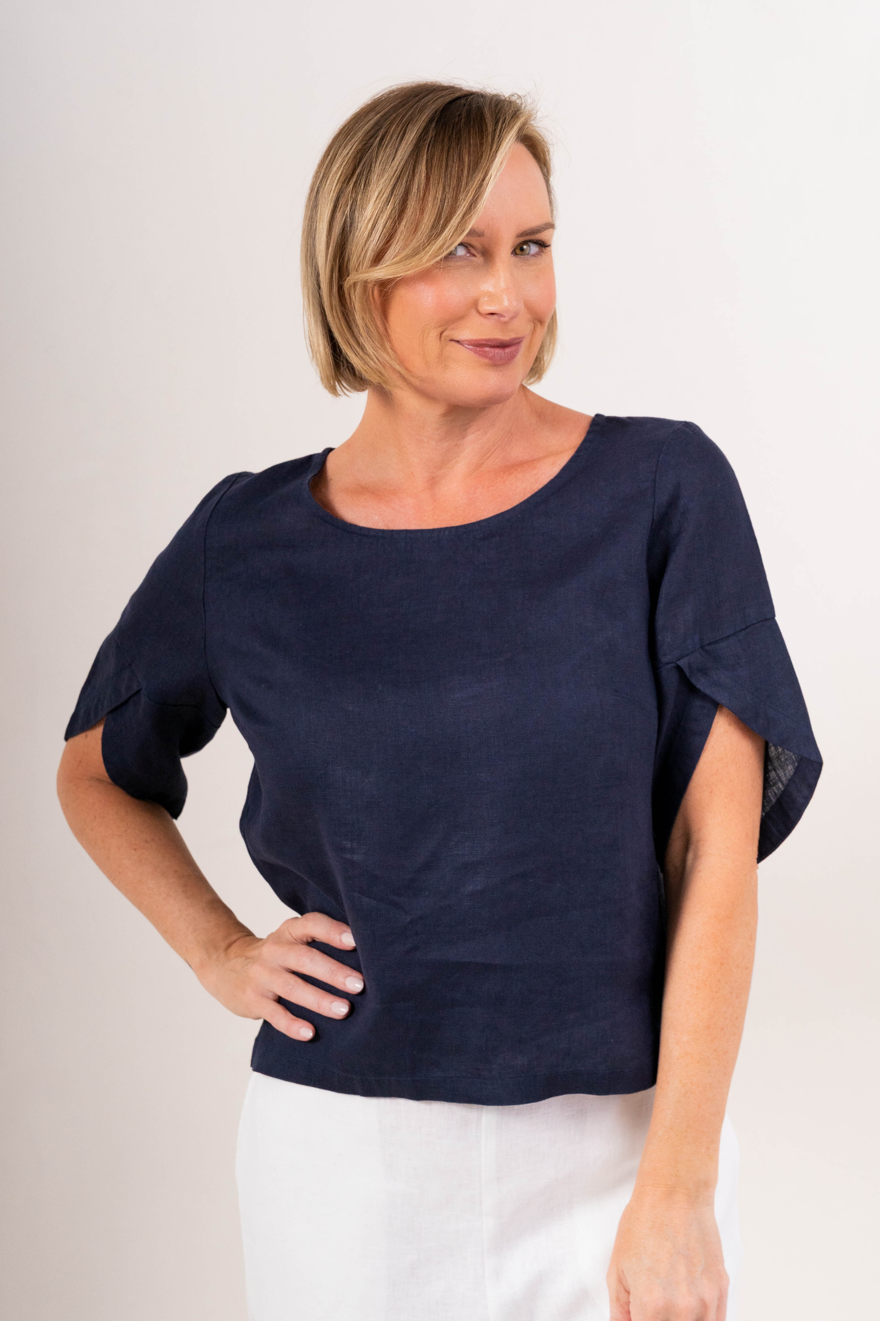 A woman wearing a navy See Saw linen short-sleeve top with ruffled or layered sleeve details and white pants, photographed against a plain light background.