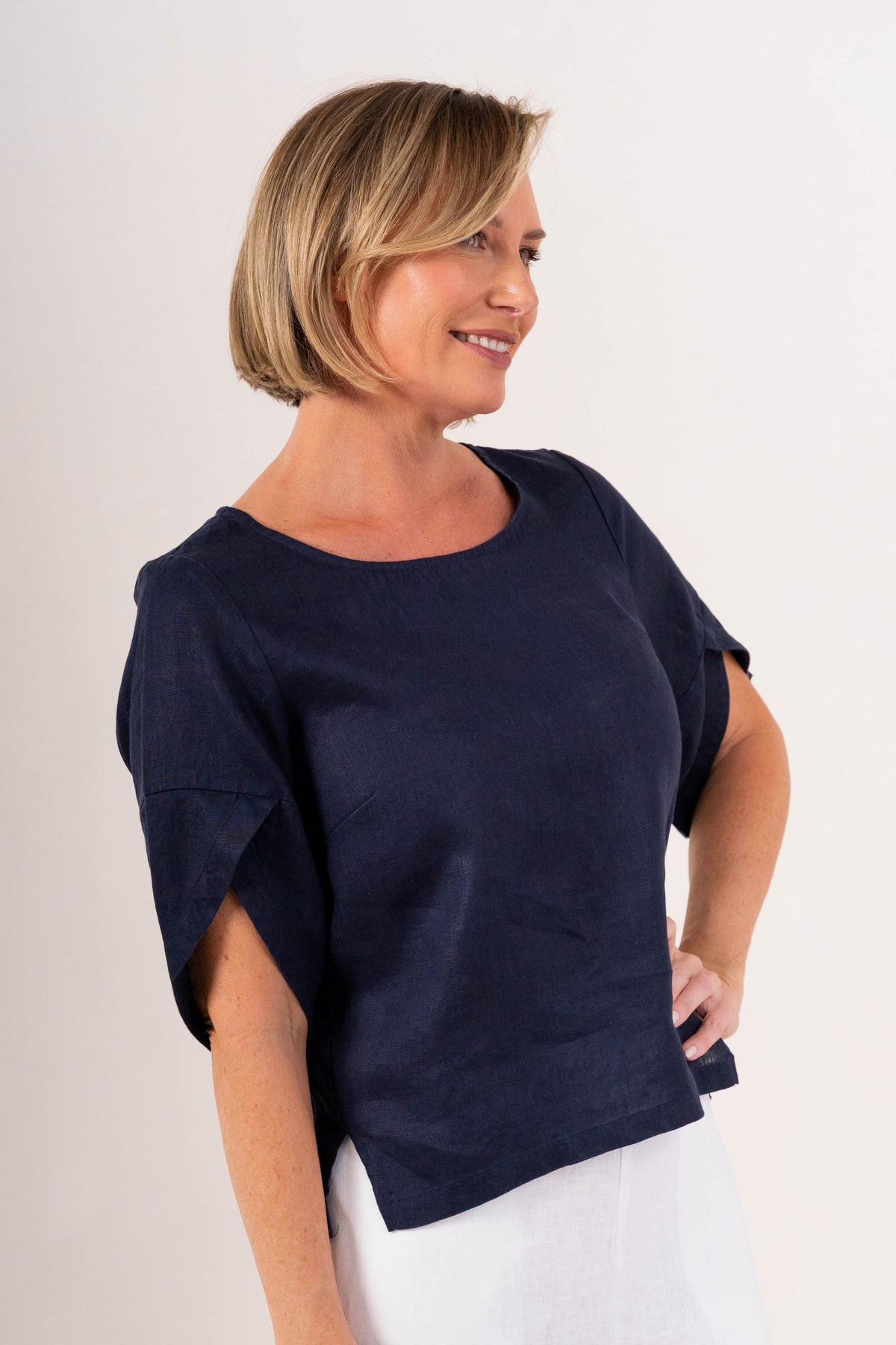 A woman wearing a navy See Saw linen short-sleeve top with ruffled or layered sleeve details and white pants, photographed against a plain light background.