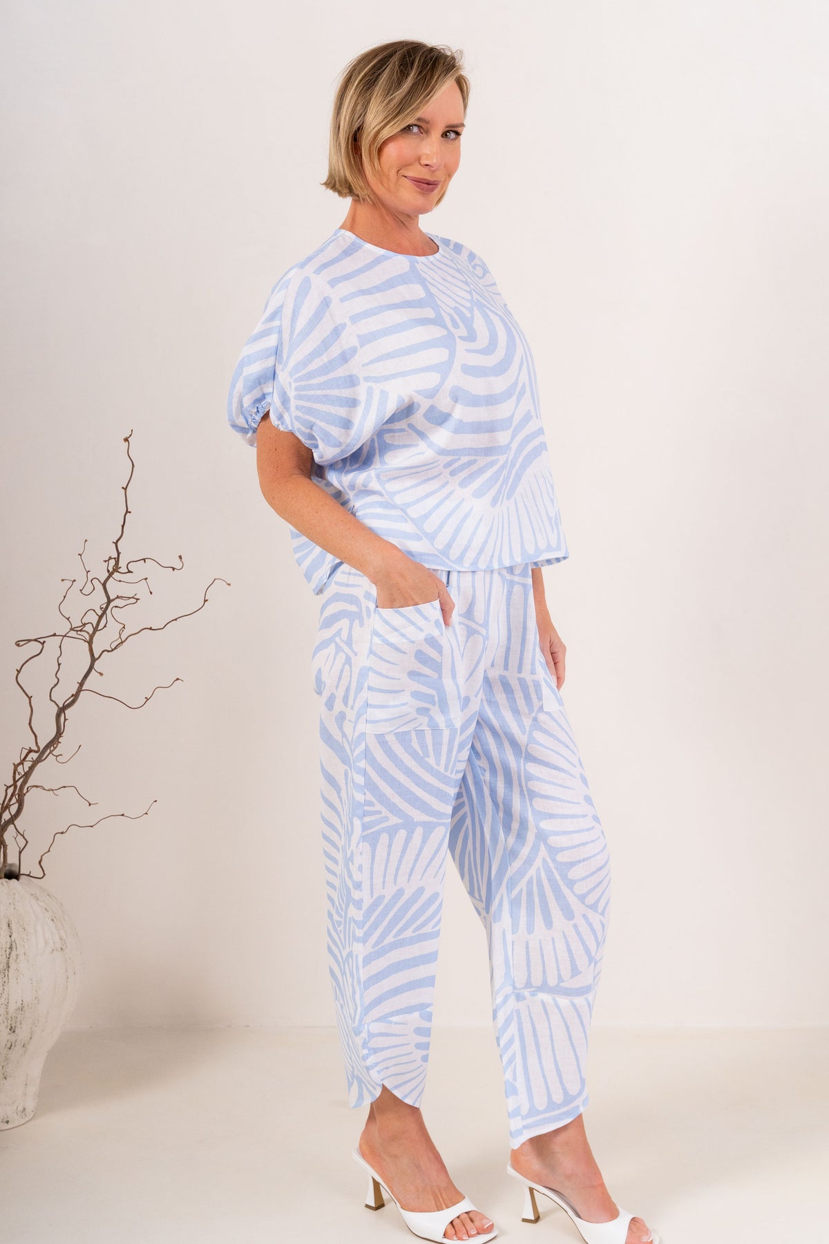 A woman modeling a Palm Breeze linen patterned two-piece set consisting of a short-sleeve top and matching pants, standing against a plain off-white studio background.