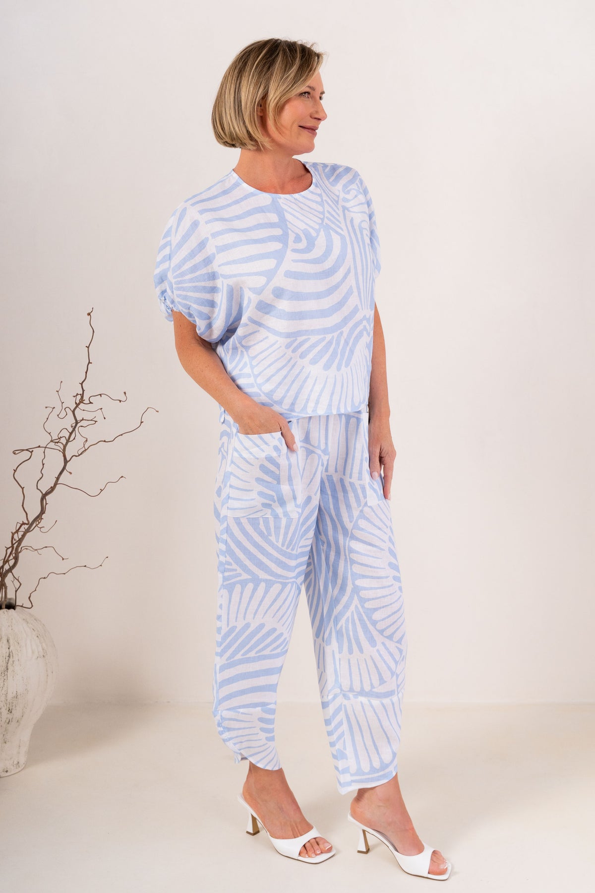 A woman modeling a light blue and white Palm Breze See Saw Linen two-piece set consisting of a short-sleeve top and matching pants, standing against a plain off-white studio background.